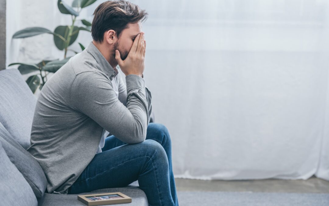 man sitting on grey couch with photo in frame and crying at home, grieving disorder concept